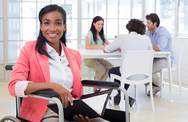 African american woman in wheelchair looks at camera while holding a folder. Colleagues sit at table in background